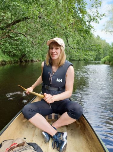Nainen hymyilee ja istuu melomassa kanootissa. Taustalla joen pinta on tasainen, mutta väreilee hieman ja joen varressa on vehreitä puita.