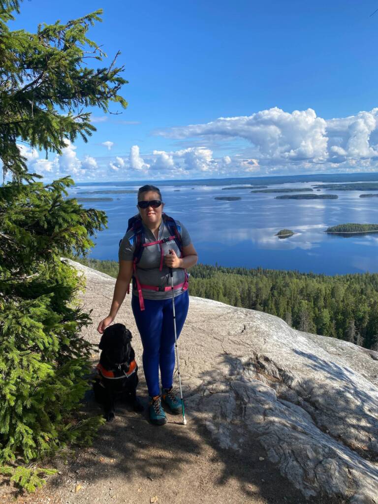 Kuva kauniista Kolin maisemasta: avautuu järvimaisema, puita, aurinkoinen päivä. Minä seison harmaassa paidassa, sinisissä trikoissa ja aurinkolasit päässä, valkoinen keppi kädessä. Vieressäni istuu Muusa, jolla päällään oranssit opaskoiraliivit.