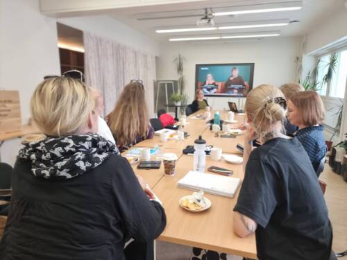 Ihmisiä istuu ruskean, pitkän pöydän ympärillä ja katsovat pöydän toisessa päässä seinällä olevaa televisiota. Huone on valkoinen ja valoisa.